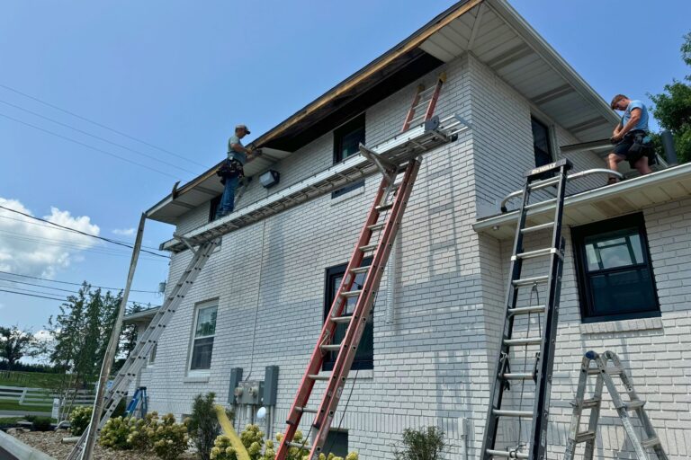 Two workers on a white brick house roof with ladders, scaffolding, and tools during daylight repair project.