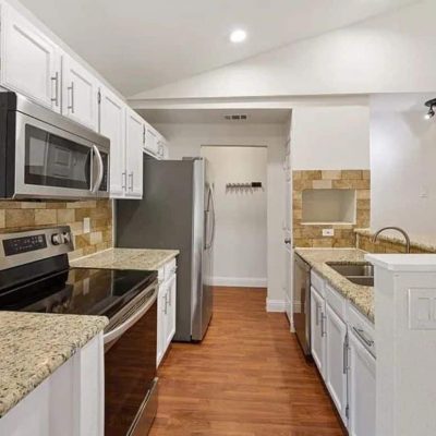Bright, modern kitchen featuring white Shaker cabinets, black countertops, brass fixtures, open shelving, stainless appliances, and warm hardwood floors